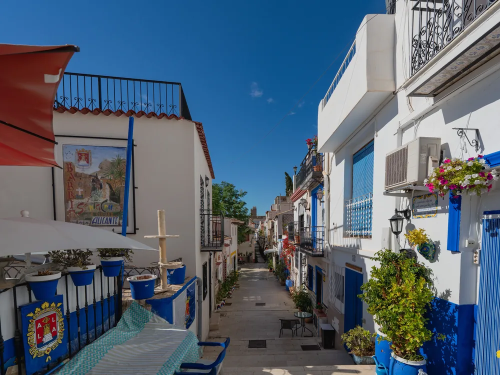 Calle empedrada del barrio de Santa Cruz de Alicante con fachadas blancas y azules y macetas con flores