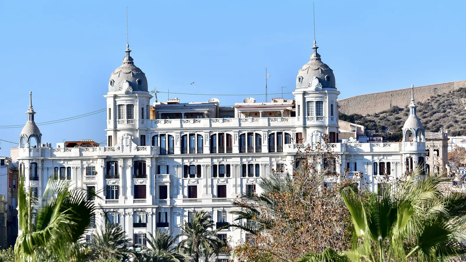 Fachada de la Casa Carbonell de Alicante entre palmeras, con el monte Benacantil al fondo