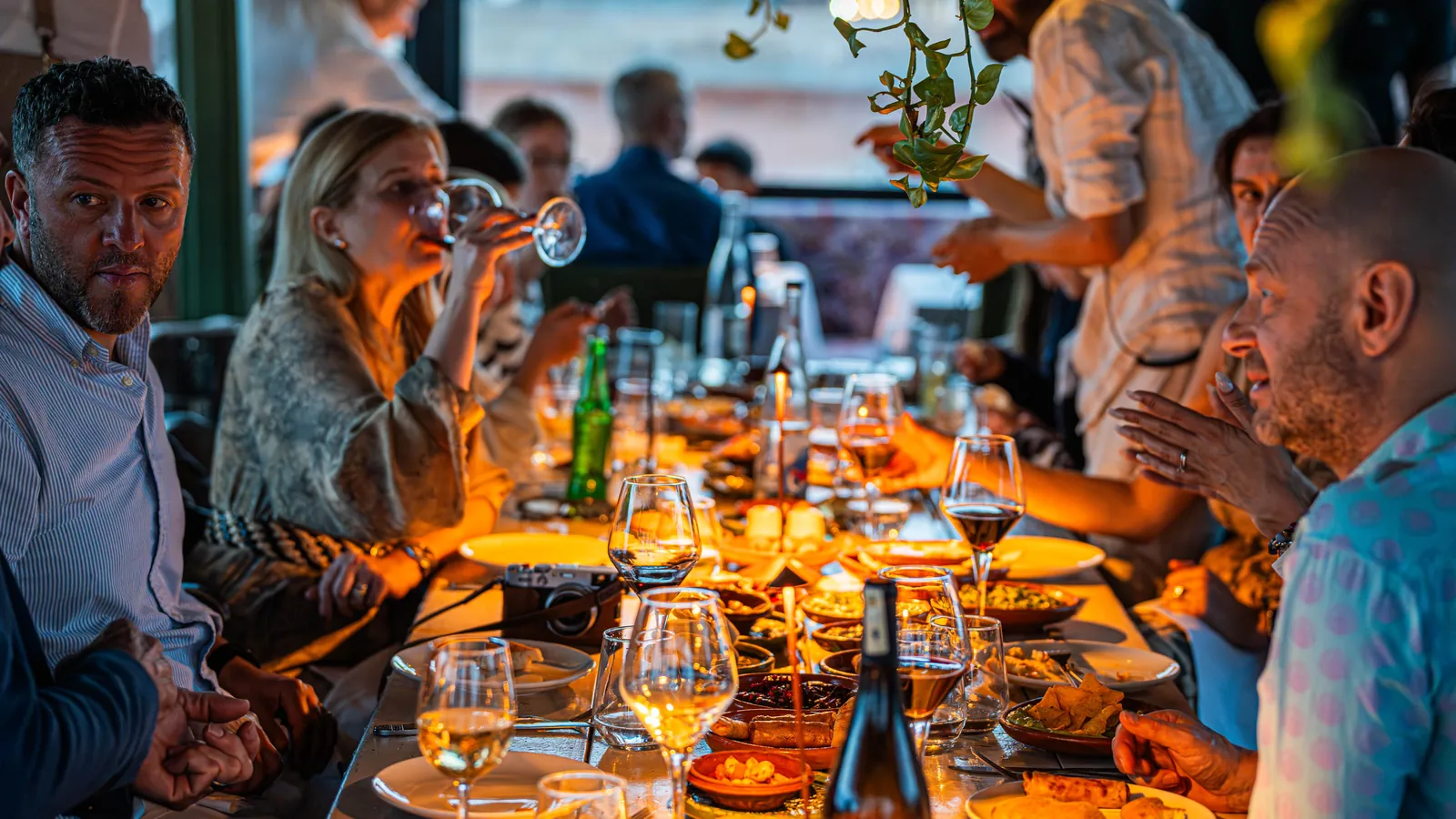 Comensales compartiendo platos y vino en una mesa larga de la terraza de un restaurante al atardecer