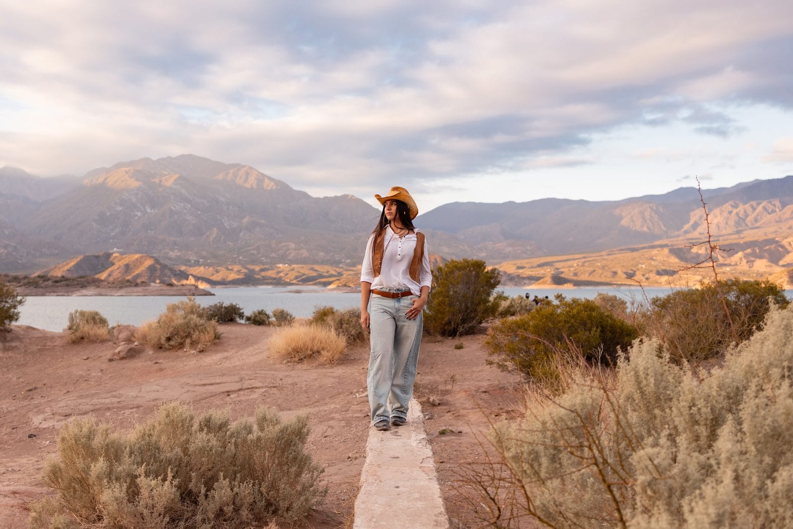 Mujer con sombrero caminando por un sendero árido junto a un embalse rodeado de montañas