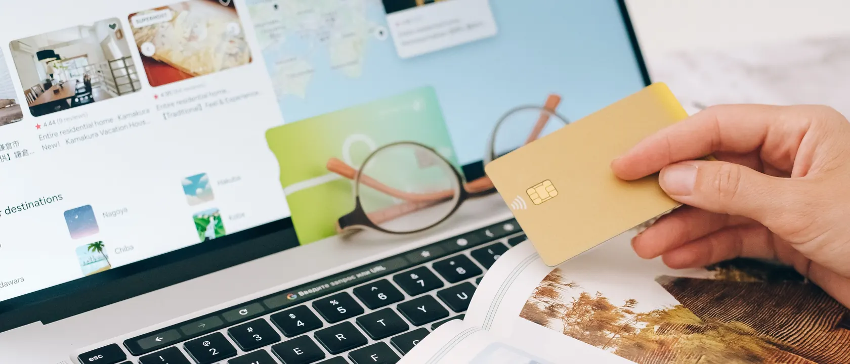 Persona sujetando una tarjeta dorada frente a un portátil con una web de alojamientos y una revista de viajes abierta