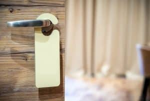 Colgador de puerta en blanco colgado del pomo metálico de una puerta de madera, con una habitación desenfocada al fondo