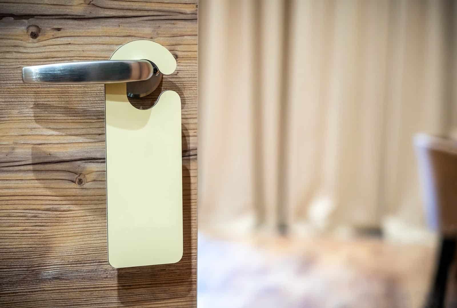 Colgador de puerta en blanco colgado del pomo metálico de una puerta de madera, con una habitación desenfocada al fondo