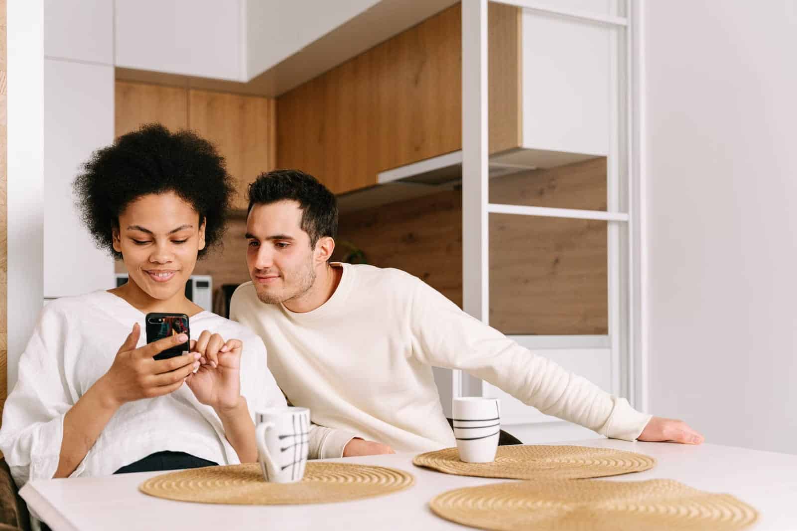Pareja sentada a la mesa de una cocina mirando juntos un teléfono móvil, con dos tazas delante