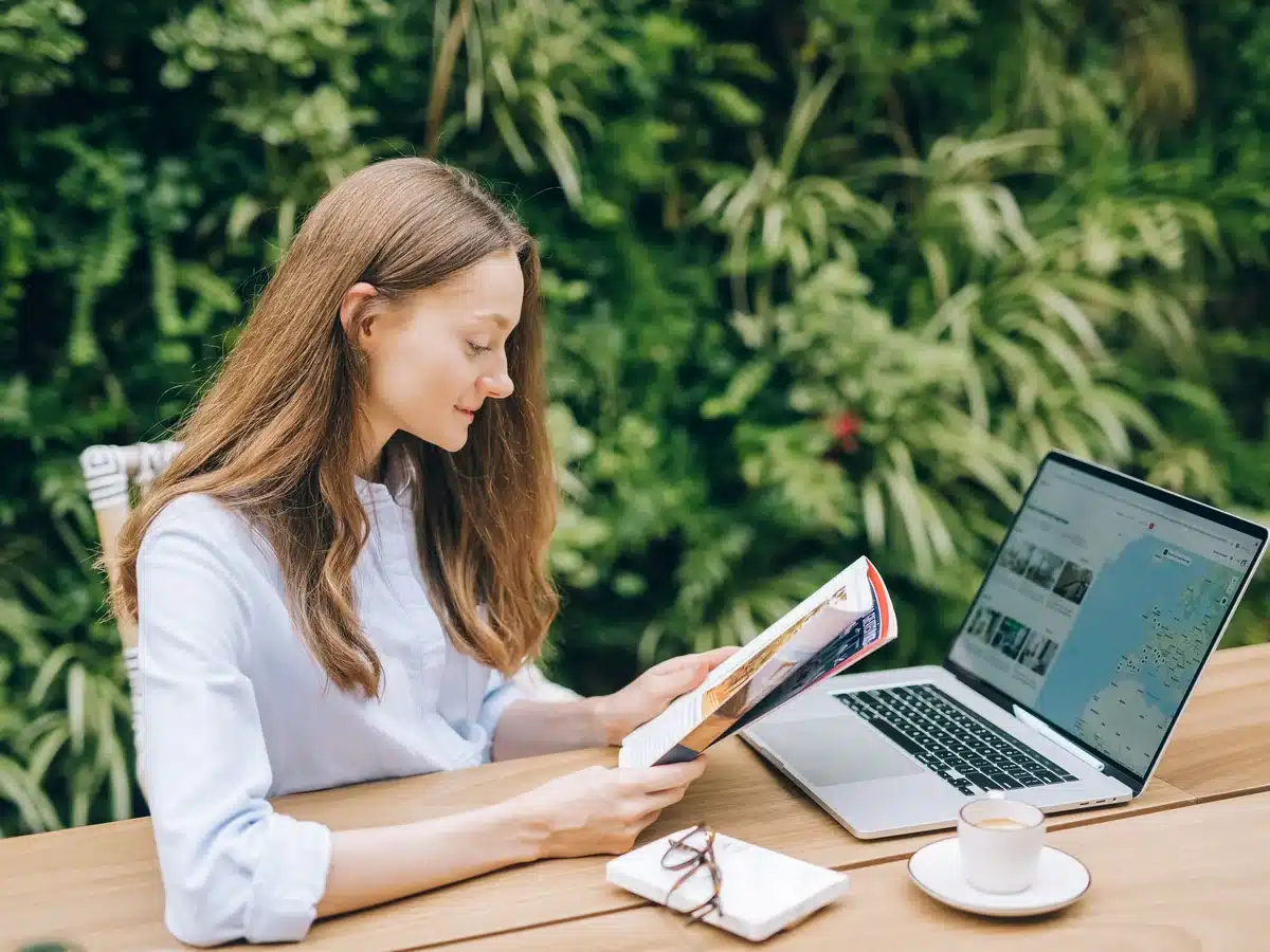 Mujer consultando una revista de viajes junto a un portátil con un buscador de alojamientos