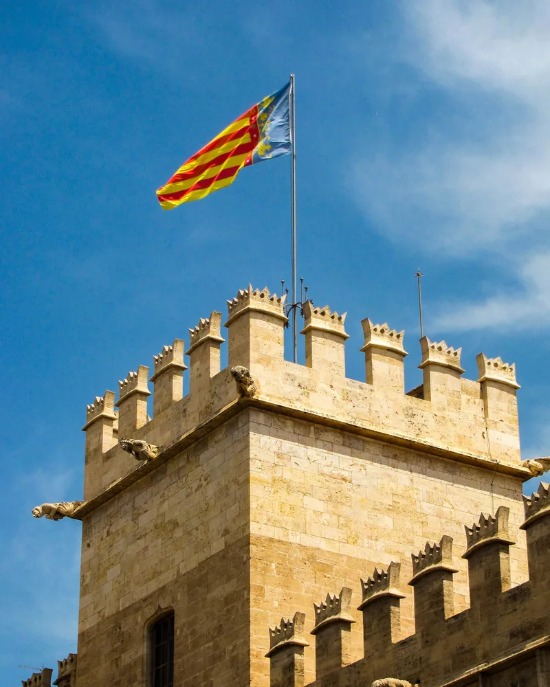 Torre gótica almenada con gárgolas y la senyera valenciana ondeando sobre el cielo azul